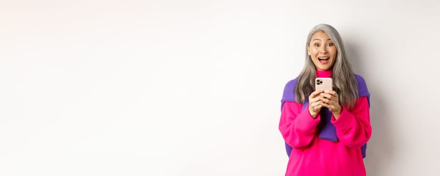 Surprised asian woman smiling at camera after reading promotion on smartphone, standing with mobile phone over white background.