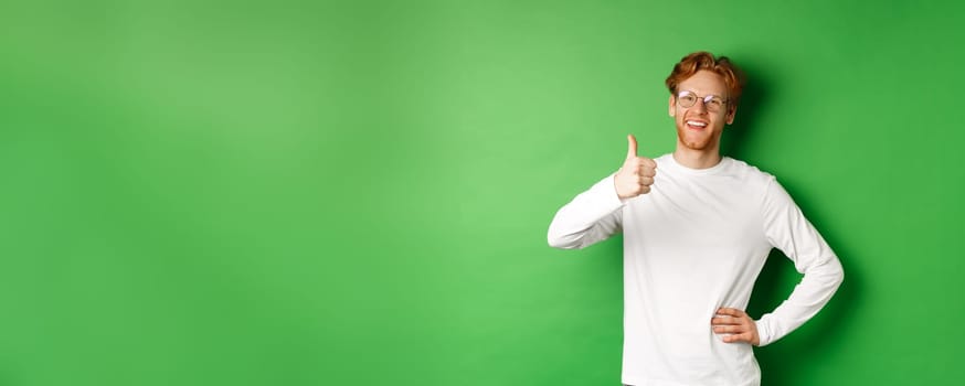 Cheerful young man with beard and red hair, wearing glasses, showing thumb-up in approval and smiling, say yes, standing over green background.