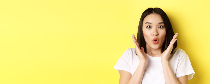 Close up of surprised asian girl say wow, stare at camera amzed with hands near face, standing over yellow background.