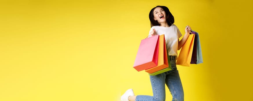 Full size shot of beautiful asian woman jumping with shopping bags and smiling happy, yellow background.