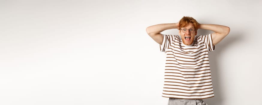 Frustrated redhead man panicking, screaming and holding hands on head, standing on white background.