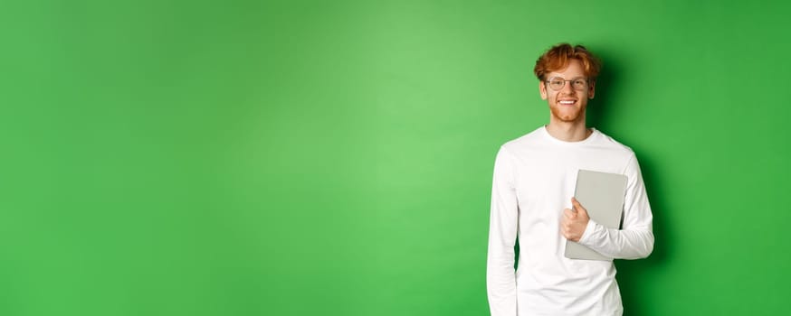 Handsome young man with red hair, wearing glasses and long-sleeve t-shirt, holding laptop on green background.