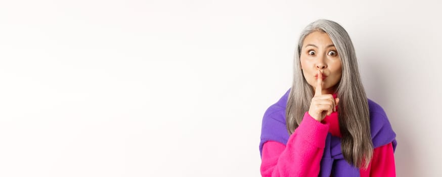 Close-up of stylish senior asian woman in hipster outfit telling hush, shushing at looking at camera, show taboo gesture, standing over white background.