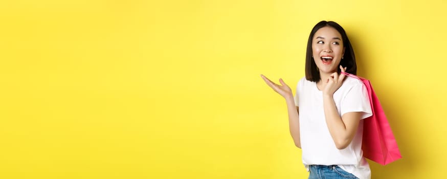 Cheerful asian female shopper looking amused, holding shopping bag and looking right, standing over yellow background.