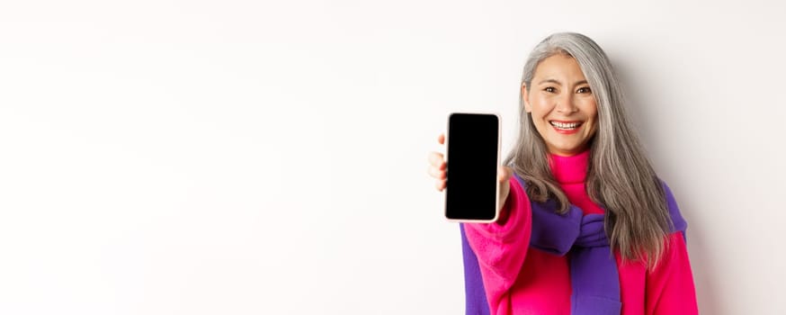 Online shopping. Close up of modern asian senior woman extending hand with mobile phone, showing blank smartphone screen and smiling, standing over white background.