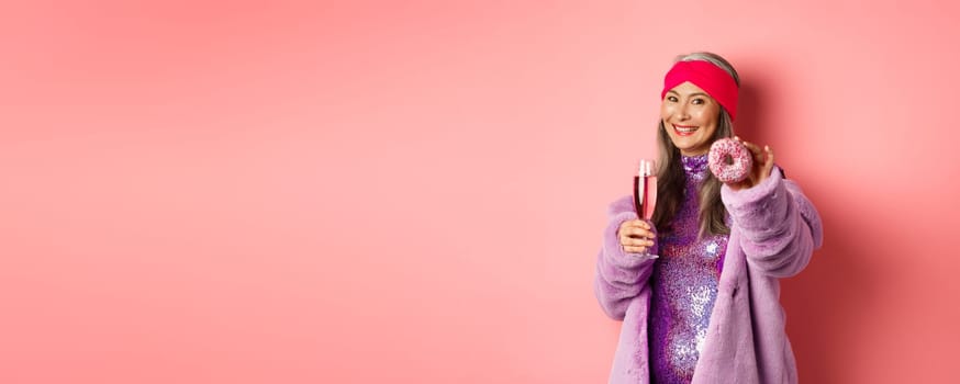 Cheerful asian elderly woman in fancy clothes drinking champagne, giving you donut, extend hand with dessert and smiling, pink background.