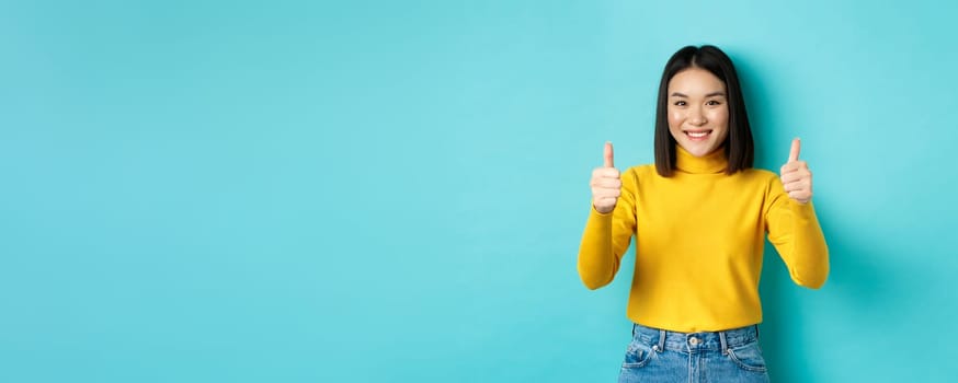 Cheerful asian female model showing thumbs up gesture, smiling and looking impressed, praise good product, standing over blue background.