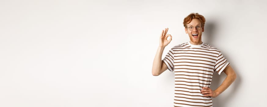 Excellent job. Happy redhead man praising you, showing OK sign and smiling amazed, approve and like something good, standing over white background.