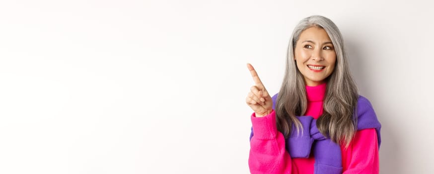 Close-up of beautiful asian senior woman smiling, looking and pointing upper left corner advertisement, checking out promo offer, standing over white background.