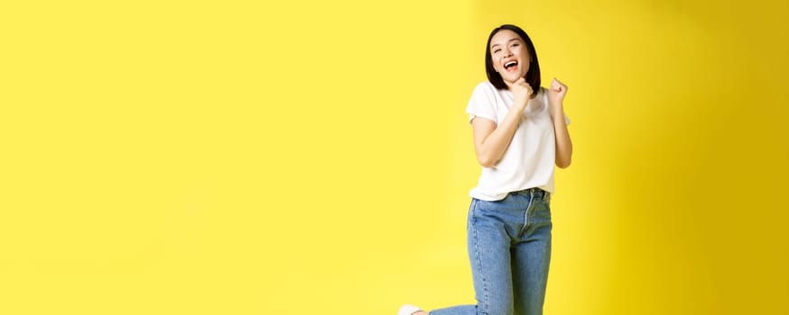 Full size shot of happy asian woman dancing and jumping from happiness, winning and celebrating victory, posing over yellow background.