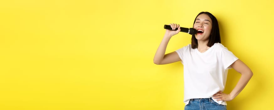 Happy asian woman singing song in karaoke, holding microphone, standing over yellow background.