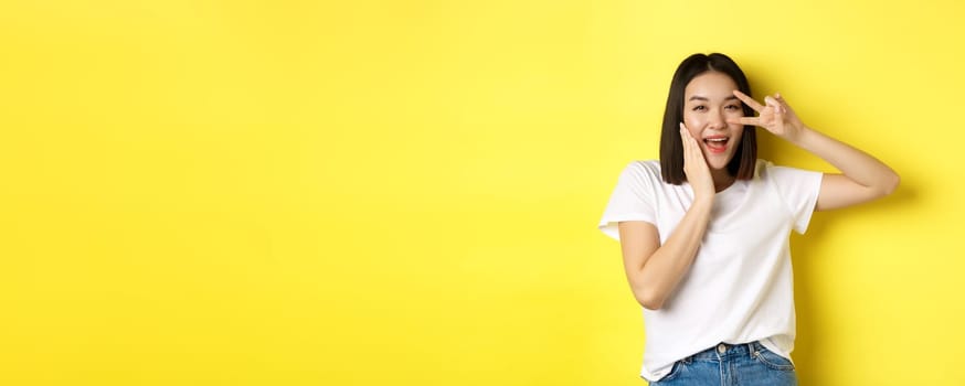 Lovely asian girl in white t-shirt posing with hand on cheek, showing peace sign on eye, standing over yellow background.