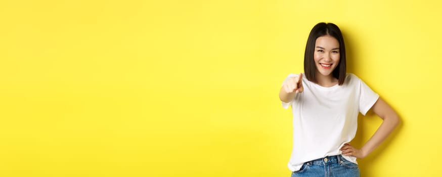 We need you. Confident asian woman smiling, pointing finger at camera, inviting to join her, beckon or choosing someone, standing over yellow background.