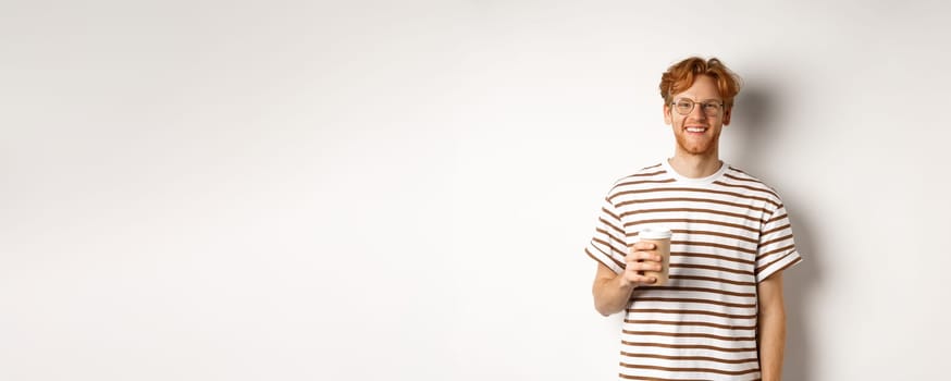 Handsome young man with beard and red messy hair, wearing glasses with striped t-shirt, drinking coffee from takeaway and smiling, white background.