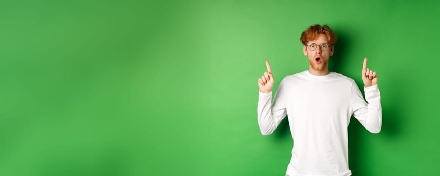 Shocked and worried redhead man in glasses gasping and staring at camera, pointing fingers up at advertisement, standing over green background.