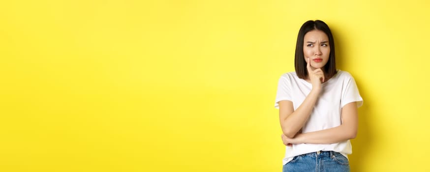 Beauty and fashion concept. Pensive asian woman thinking, looking thoughtful while pondering something, standing over yellow background.