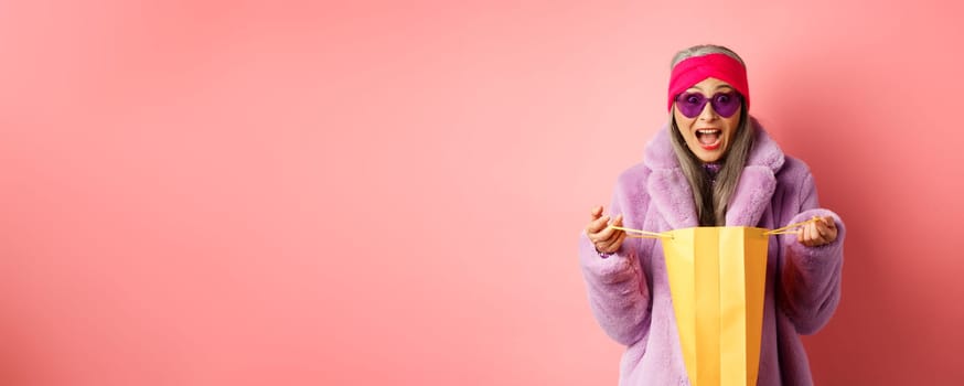 Shopping and fashion concept. Stylish asian elderly woman in sunglasses and faux fur coat open paper bag with gifts, looking amazed at camera, pink background.