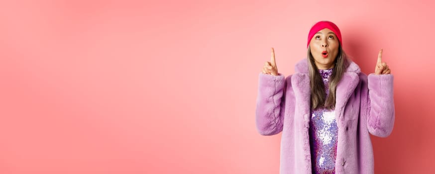 Impressed asian elderly woman in stylish fake-fur coat checking out promo, looking and pointing fingers up, standing over pink background.