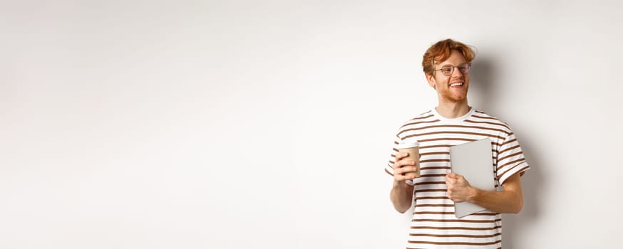 Handsome redhead male employee in glasses having break, drinking coffee and holding laptop, standing over white background.