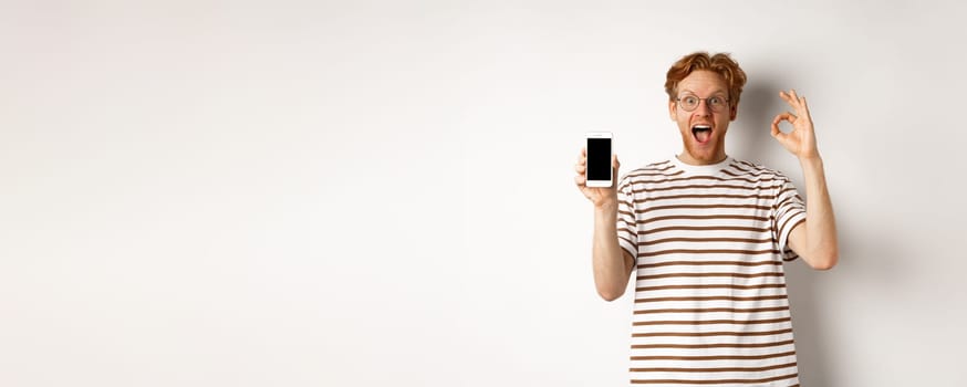 Technology and e-commerce concept. Young man with red hair showing okay sign and blank smartphone screen, praising awesome app, standing over white background.