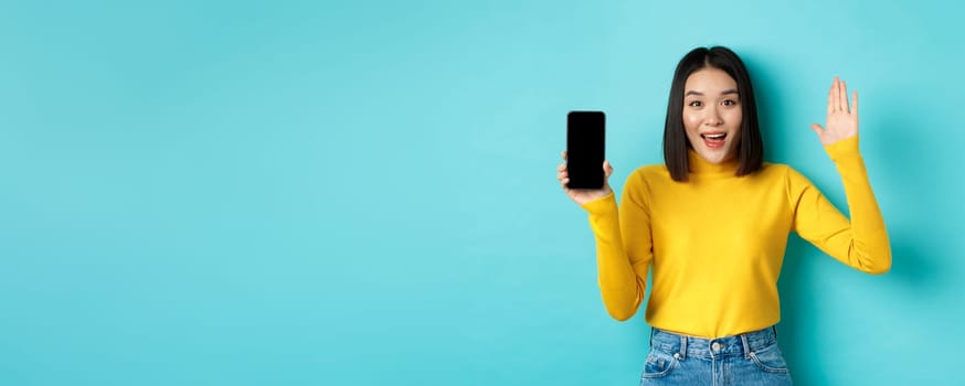 Cheerful asian woman showing empty smartphone screen and raising hand to say hello, greeting you, standing against blue background.