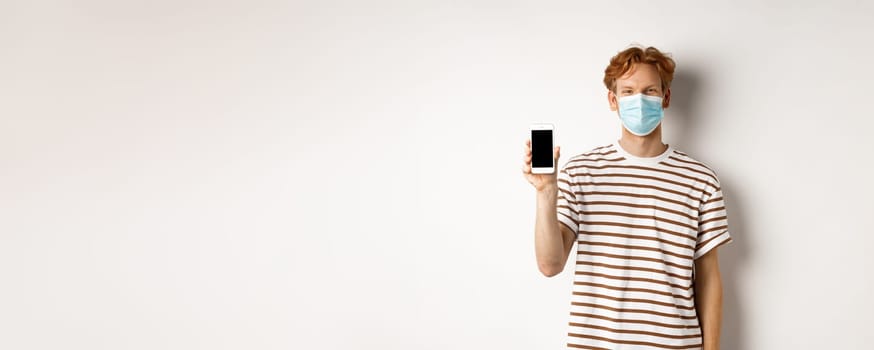 Covid-19, pandemic and social distancing concept. Handsome young man with face mask, showing black smartphone screen and smiling, standing over white background.