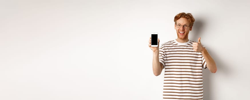 Technology and e-commerce concept. Young man with red hair showing thumbs-up and blank smartphone screen, recommending app, white background.