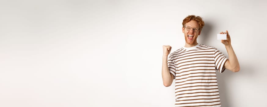 Shopping and finance concept. Young man winning bank prize, showing plastic credit card and making fist pump, screaming from joy and satisfaction, white background.