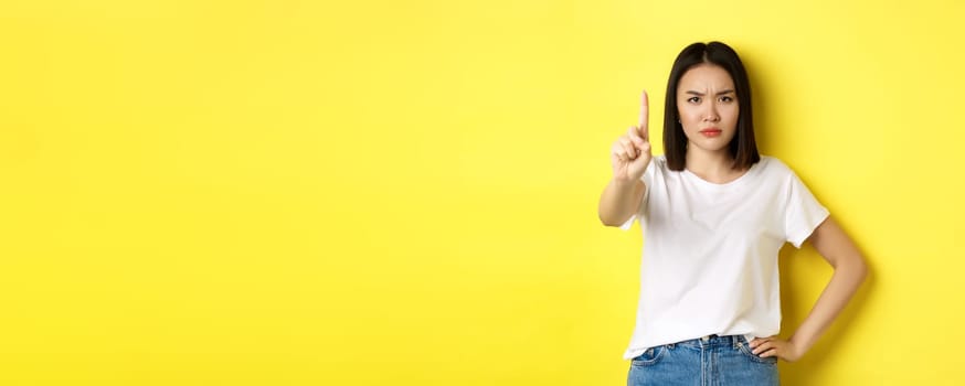 Confident and serious woman tell no, showing extended finger to stop and prohibit something bad, frowning and looking at camera self-assured, standing over yellow background.