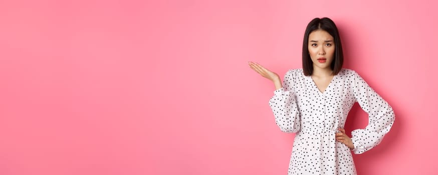 Careless asian woman raising hand and shrugging, staring at camera unbothered and questioned, so what gesture, standing over pink background.