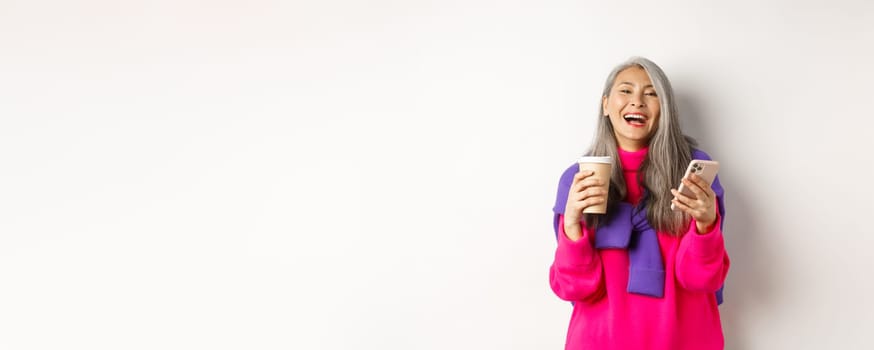 Social networking. Happy asian senior woman drinking coffee and holding smartphone, laughing at camera, standing over white background.