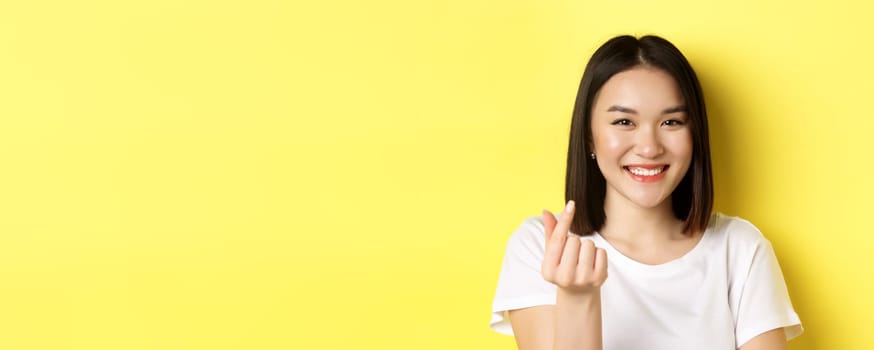 Valentines day and women concept. Close up of pretty asian girl in white t-shirt, smiling and showing finger heart, standing over yellow background.