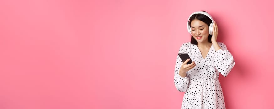 Cute asian girl listening music on headphones, looking at mobile phone and smiling, standing in dress against pink background.