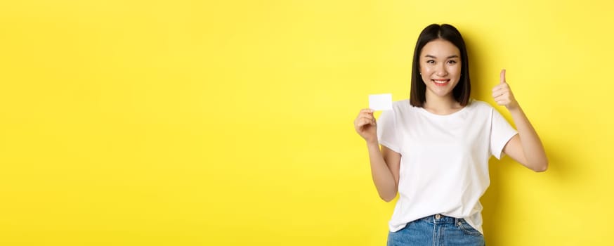 Young asian woman in casual white t-shirt showing plastic credit card and thumb up gesture, approve and recommend, smiling at camera, yellow background.