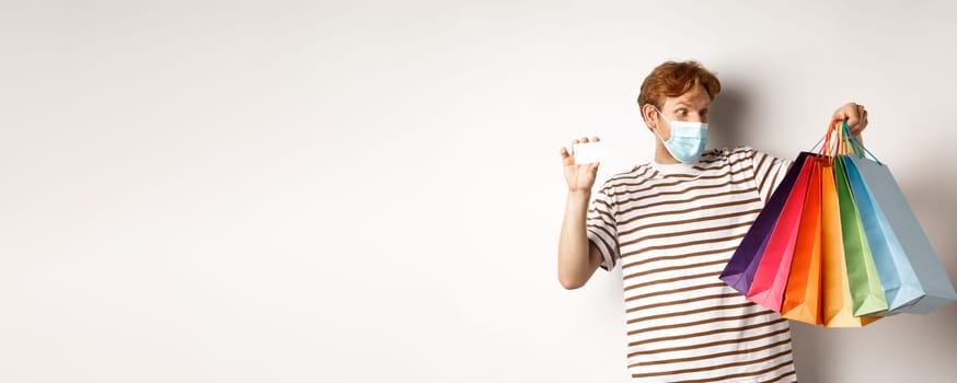 Covid, quarantine and lifestyle concept. Amazed redhead man looking at shopping bags after buying with plastic credit card of bank, white background.