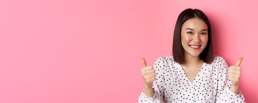 Beauty and lifestyle concept. Close-up of cute asian woman showing support, make thumbs-up gesture and smiling, like and praise you, standing over pink background.