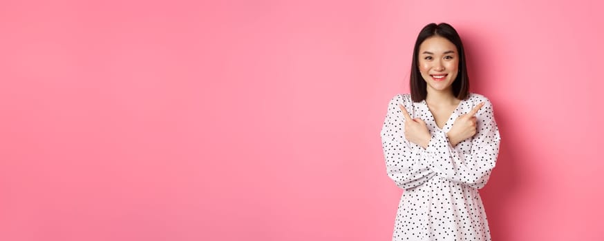 Beautiful asian girl in romantic dress pointing fingers sideways, showing two variants on shopping, smiling at camera, standing against pink background.