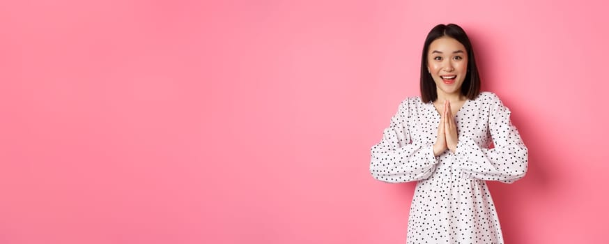 Beautiful asian woman thanking you, holding hands together in appreciation gesture, smiling happy at camera, standing grateful over pink background.