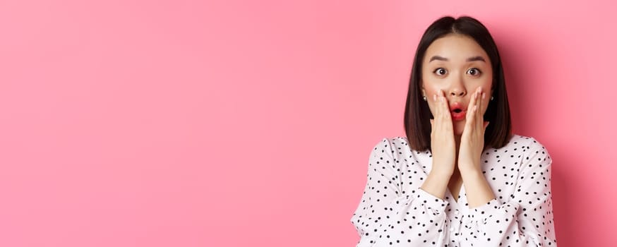 Close-up of surprised and amazed asian woman staring at camera, gasping impressed, holding hands near mouth, standing over pink background.