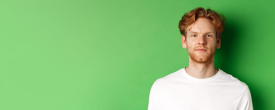 Close-up of young man with red messy hair and beard looking at camera, standing over green background.