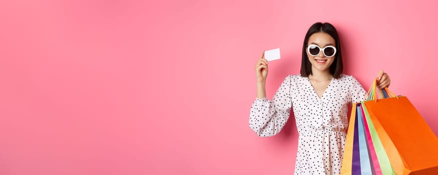 Beautiful asian woman in sunglasses going shopping, holding bags and showing credit card, standing over pink background.