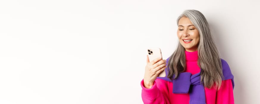 Close-up of fashionable asian senior woman looking at smartphone and reading message, smiling at mobile screen, standing over white background.