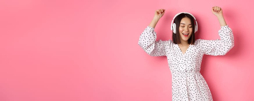 Happy young asian woman dancing and having fun, listening music in headphones, standing over pink background.