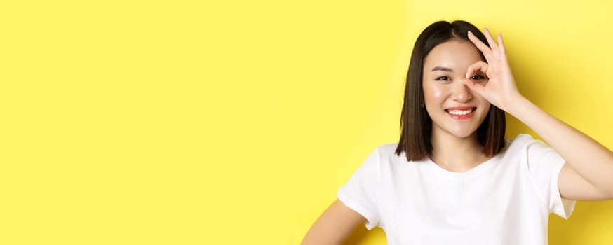 Close up of cute asian girl feeling happy, showing OK sign on eye and smiling, standing over yellow background.