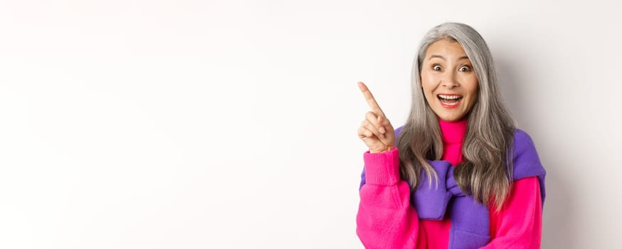 Close up of beautiful asian female model pointing finger at upper left corner, smiling and looking happy at camera, showing promo deal, white background.