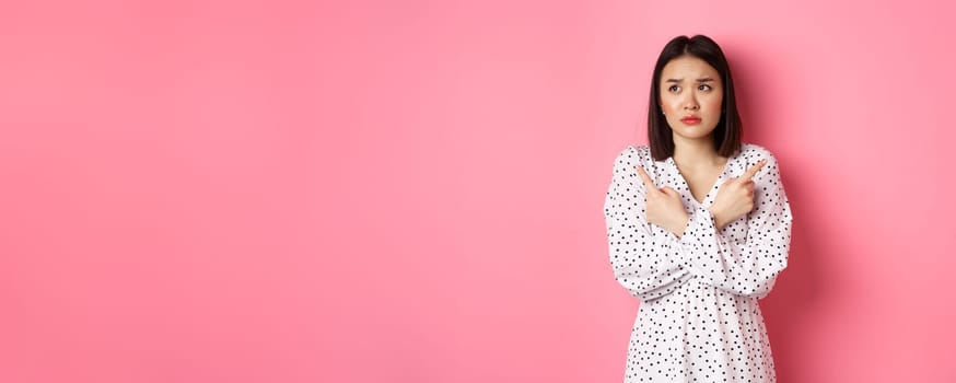 Concerned asian woman having doubts, pointing sideways and looking left with hesitant and sad face, need help with choice, standing over pink background.