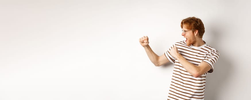 Profile of funny young man with red hair and beard, raising fists for fight, shadow boxing and shouting, standing over white background.