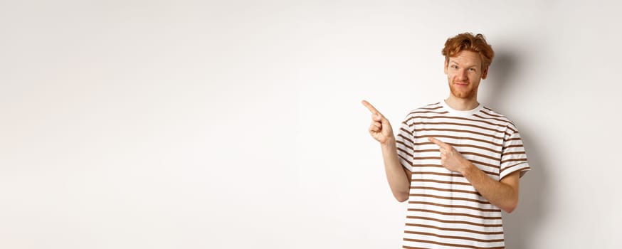 Doubtful redhead guy pointing fingers left at looking skeptical at camera, standing over white background. Copy space