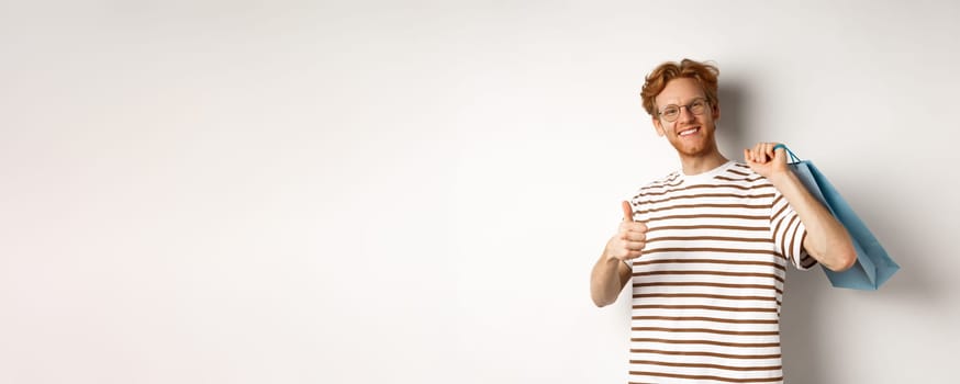 Satisfied young man leave positive review after shopping, showing thumb-up in approval and smiling, holding paper bag over shoulder, white background.