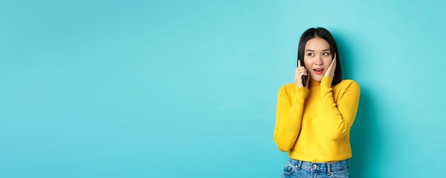 Woman looking intrigued while talking on mobile phone, receive interesting offer during call, standing over blue background.